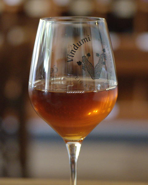 Glas Orange Wine vor dem Weinregal bei Vindumi